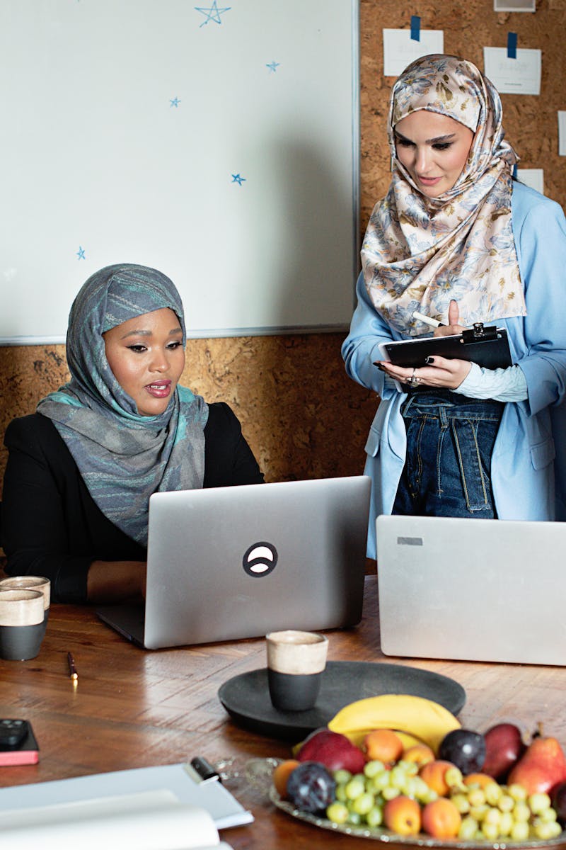 Four Muslim businesswomen in hijabs working together with laptops in a modern office setting.