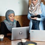 Four Muslim businesswomen in hijabs working together with laptops in a modern office setting.