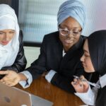 Three multicultural businesswomen in hijabs discuss work around a laptop in an office setting.
